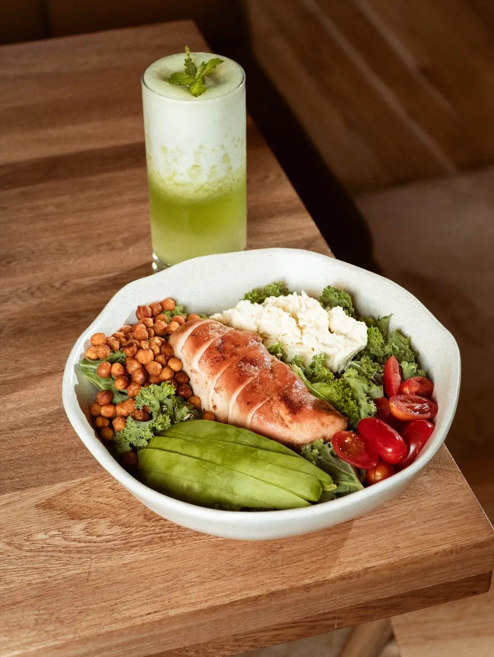 Bowl con pollo, garbanzos, aguacate y verduras, con una bebida verde al fondo