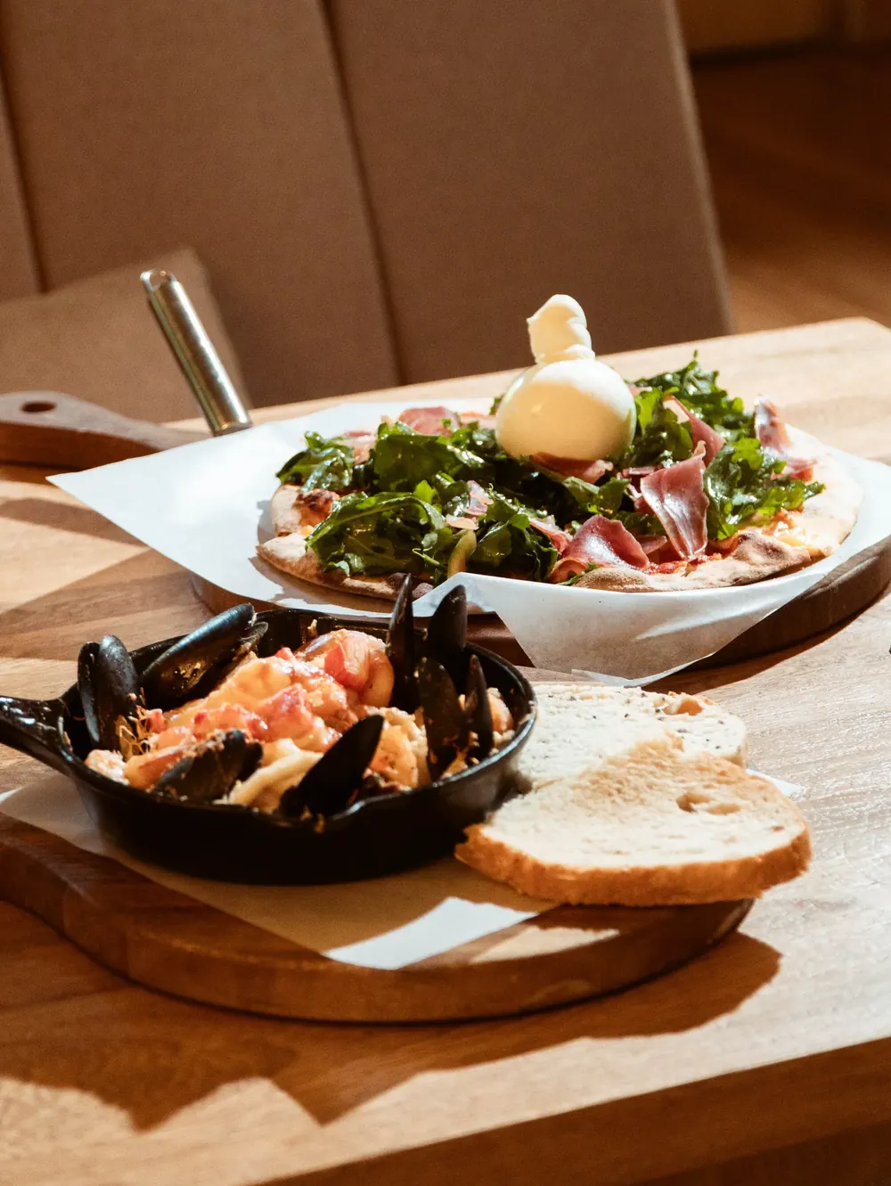 Pizza con rúgula y jamón junto a una cazuela de mariscos, sobre una mesa de madera